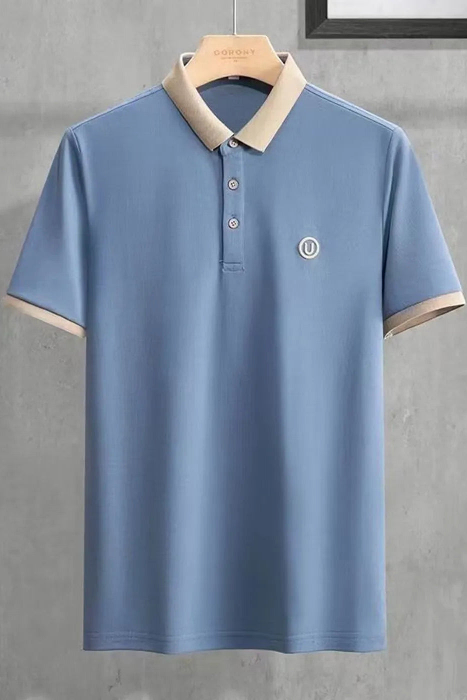 Blue polo shirt with beige collar and trim on a hanger against a gray concrete wall.