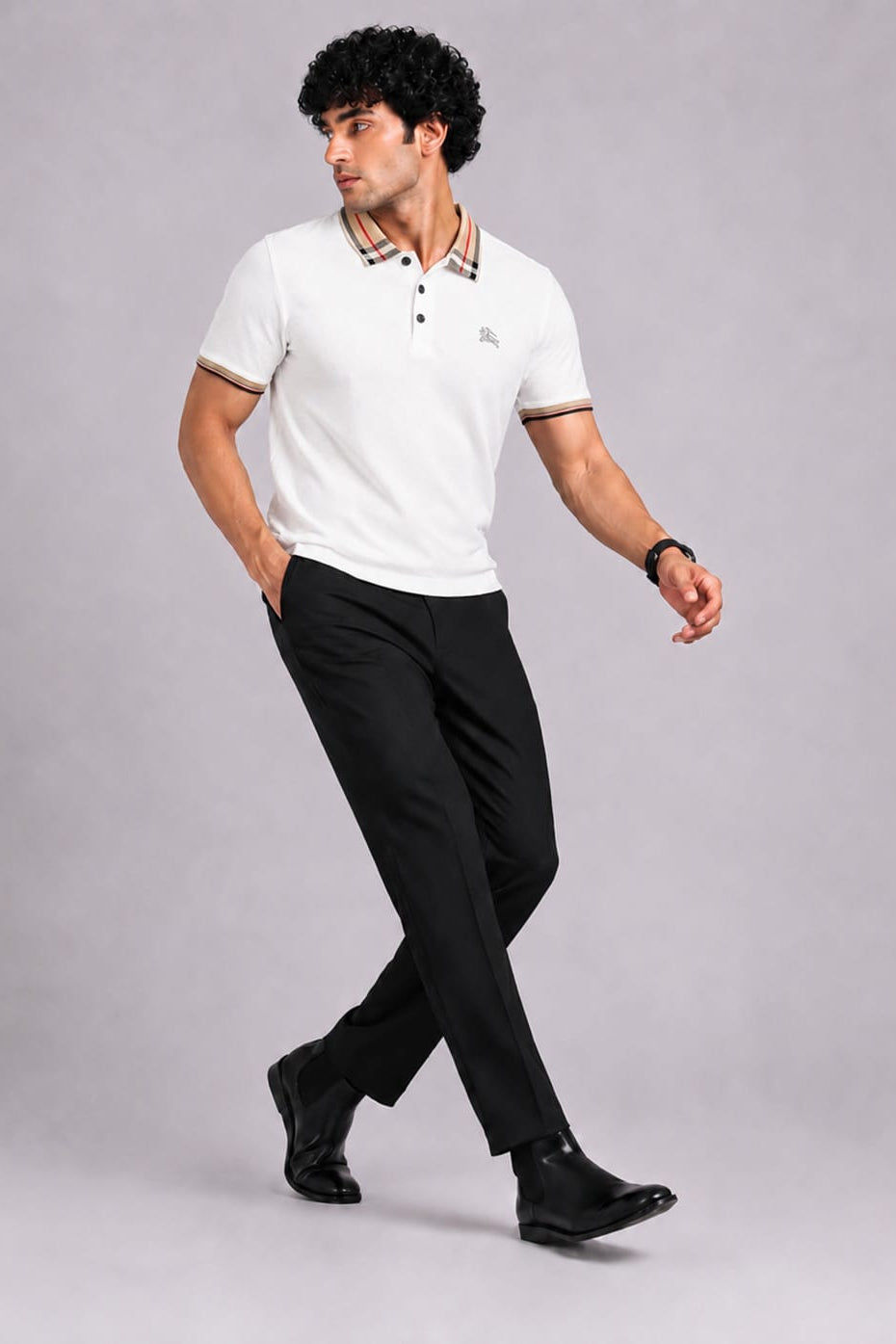 A man wearing a white short-sleeve polo shirt with a patterned collar and black trousers, walking against a solid gray studio background.