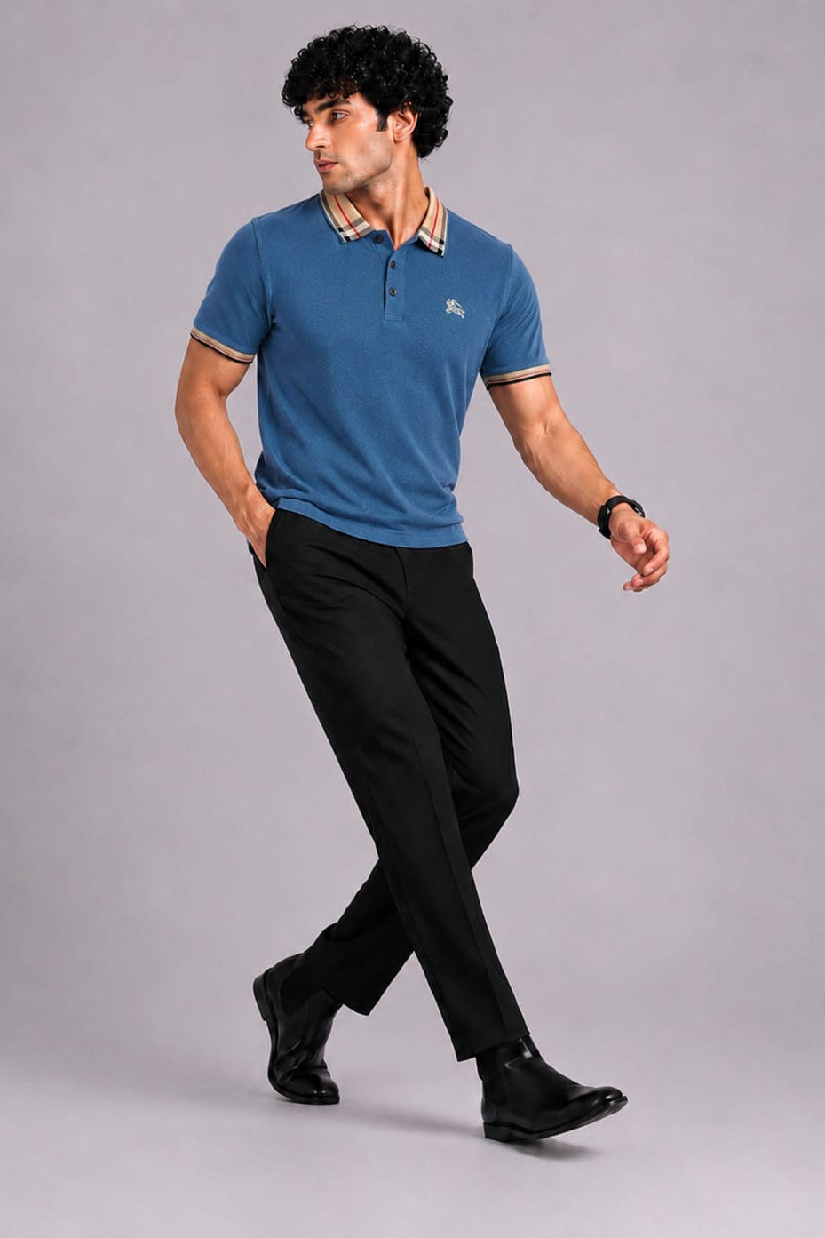 A man wearing a blue short-sleeve polo shirt with a beige and black striped collar, black trousers, and black shoes, walking against a solid gray studio background.