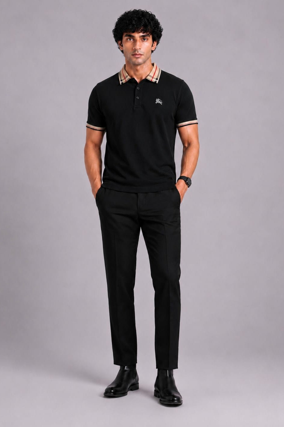 A man modeling a black short-sleeve polo shirt with a beige and white patterned collar and sleeve trim, paired with black trousers and black dress shoes, against a solid gray studio background.