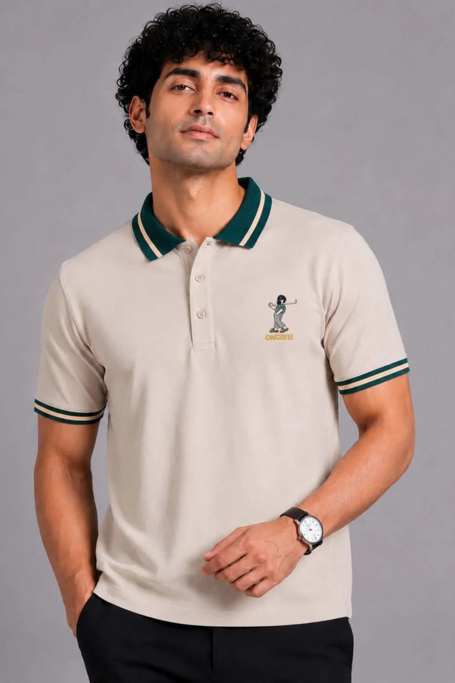 A man wearing a beige short-sleeve polo shirt with a dark green and white striped collar and sleeve cuffs, featuring a small embroidered cricket player logo on the left chest.