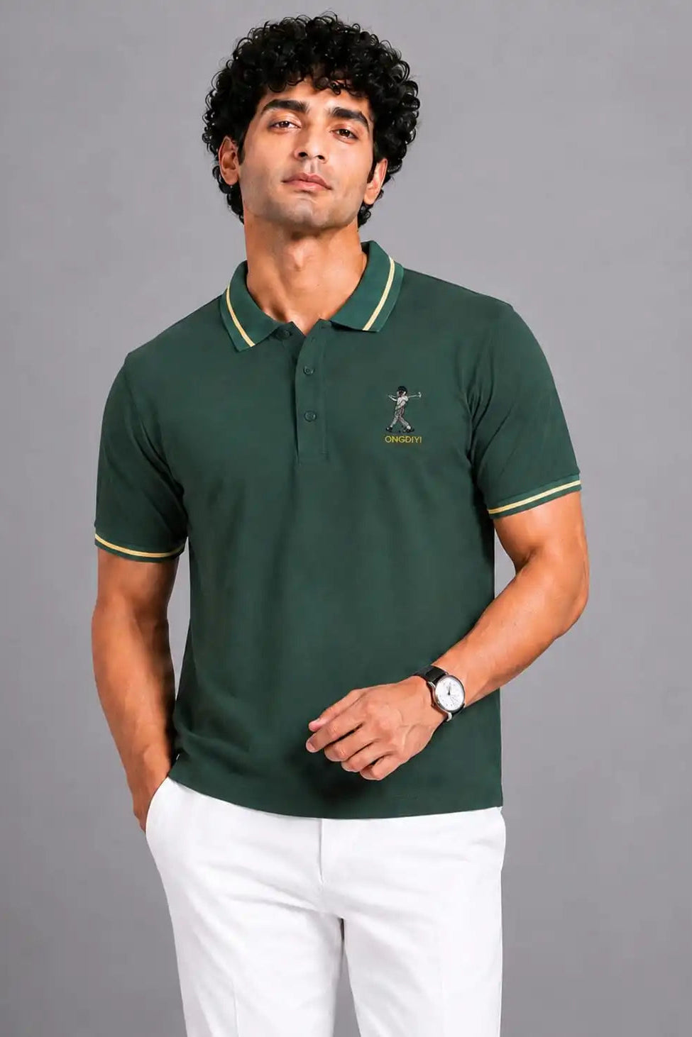 A man wearing a dark green polo shirt with light green and white striped collar and sleeve cuffs, paired with white trousers, standing against a solid gray studio background.